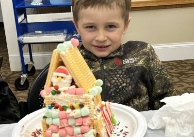 A smiling young boy showcases his handcrafted gingerbread house, filled with colorful decorations.