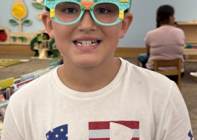 Child smiling while wearing turquoise LEGO glasses and a white T-shirt with a patterned logo.