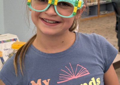 A young person wearing LEGO glasses and a blue t-shirt stands in the library.