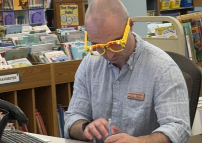 Man wearing LEGO glasses in a library, seated at a desk with books in the background.