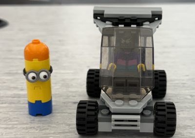 A LEGO vehicle with a minifigure and a yellow capsule-shaped toy with goggles on a gray surface.