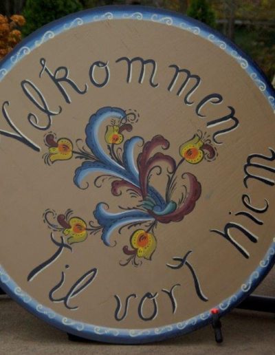 The image shows a large, circular wooden sign with decorative patterns and text in a script font. The sign has a beige background with a blue border featuring white wave-like patterns. In the center, there is an intricate floral design with swirling blue and burgundy leaves and yellow flowers. The text is inscribed around the top and bottom edges of the circle in black script. The sign appears to be positioned outdoors on a patio with a grassy area and orange-brown bushes in the background, adjacent to a brown building.