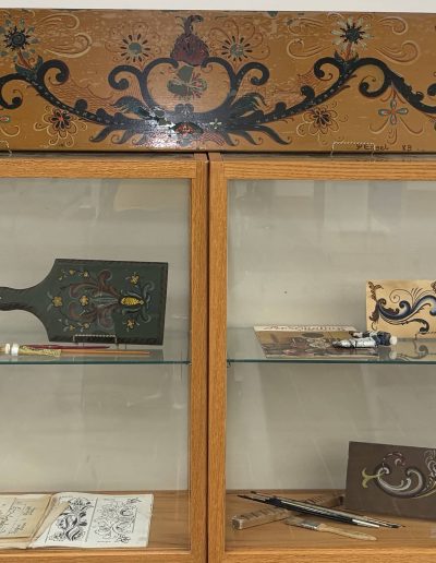 The image shows a wooden cabinet with two shelves displaying various items related to Norwegian rosemaling. On the top of the cabinet, there is a decorative wooden panel featuring an intricate floral and scroll design in earthy tones of brown, orange, and green. Beside it, there is an informational plaque titled "Norwegian Rosemaling by Peter Engel," including a picture of an artist and accompanying text. The upper shelf contains a paddle-shaped object with similar decorative patterns, some art tools like paint tubes, and a small rectangular box. The lower shelf showcases sketches and patterns of floral designs on paper along with brushes and other painting tools. The display highlights traditional Norwegian decorative painting, showcasing items likely used by an artist in creating rosemaling art.