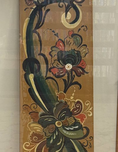 The image features a vertically oriented, rectangular wooden panel adorned with intricate, colorful floral patterns. The background is a warm brown hue, against which bold designs in dark green, red, cream, and black swirl in a lively manner. These floral motifs exhibit an ornate style, with swirling leaves and blooming flowers running diagonally across the panel. Each flower is distinct, characterized by layered petals and decorative flourishes. Above the panel is a glass shelf on which sits a magazine or advertisement featuring similarly ornate objects like a decorated box, a circular tray, and a small piece of furniture.