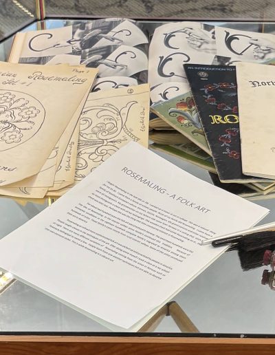 The image showcases a display of rosemaling materials under a glass case. Several booklets and sheets of paper are arranged on the display surface. On the left, a stack of papers features a drawing of a floral design with the heading "Norwegian Rosemaling" and the name "Elizabeth Bradley." To the right, several booklets with decorative covers and titles like "Traditional Norwegian Rosemaling" and "Rosemaling" are visible. In the foreground, a white sheet of paper titled "ROSEMALING - A FOLK ART" is partially readable. Near the paper are two paintbrushes with wooden handles and tubes of paint.