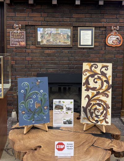 The image shows an art display against a brick wall, featuring three panels with intricate, swirling botanical designs. Each panel is placed on an easel, with two panels resting on wooden bases and one inside a glass case. The panels feature ornate floral and vine motifs in various colors: one is green with yellow and white flowers, another is blue with earthy tones, and the third is beige with prominent brown and golden swirls. Two wooden cabinets with glass tops flank the center of the display, each containing additional artworks and a framed piece of text. Behind the display, framed pictures and decorative autumn-themed signs hang on the brick wall. A wooden slab serves as the base for the central easels, with a sign that reads "STOP PLEASE DO NOT TOUCH."